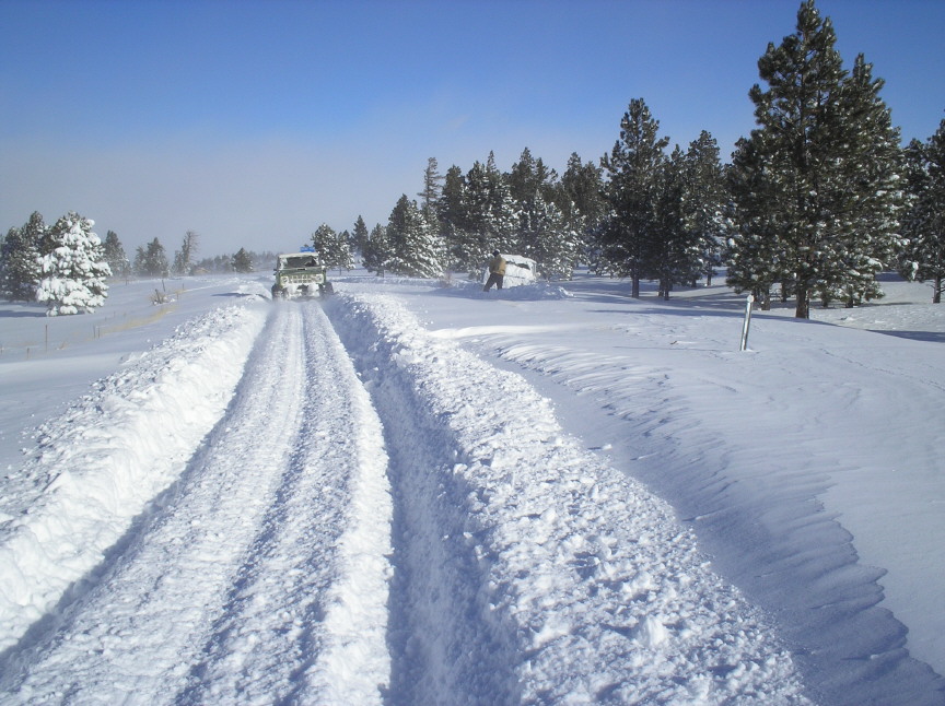 Tire tracks cut through deep snow on a forest road with a vehicle stopped in the distance