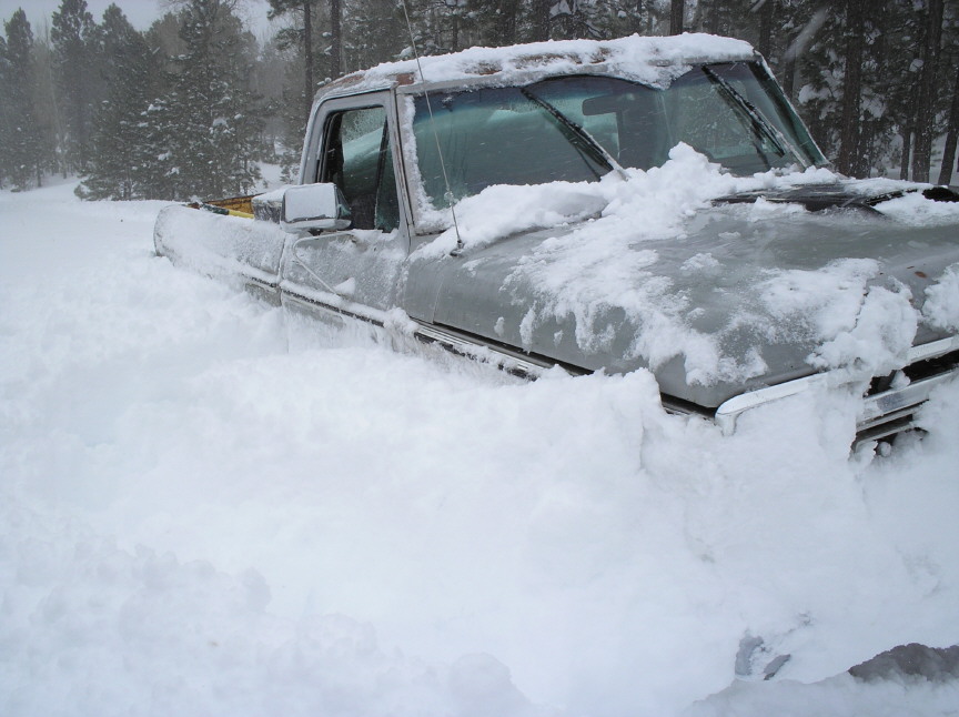 Older pickup truck stuck in deep snow with snow heaped over its hood during snowfall