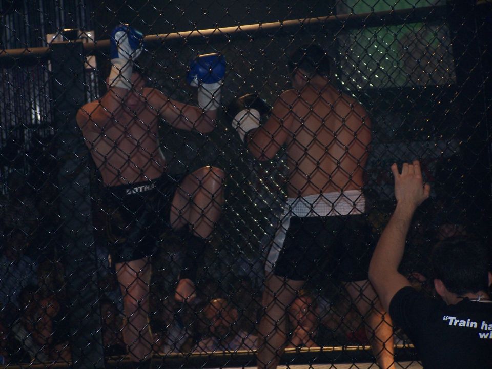 Two MMA fighters in a cage, one kicking, viewed through a chain-link fence