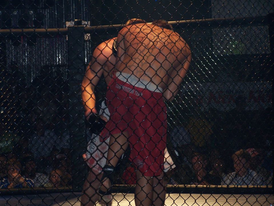 Two MMA fighters grappling inside a cage, viewed through a chain-link fence