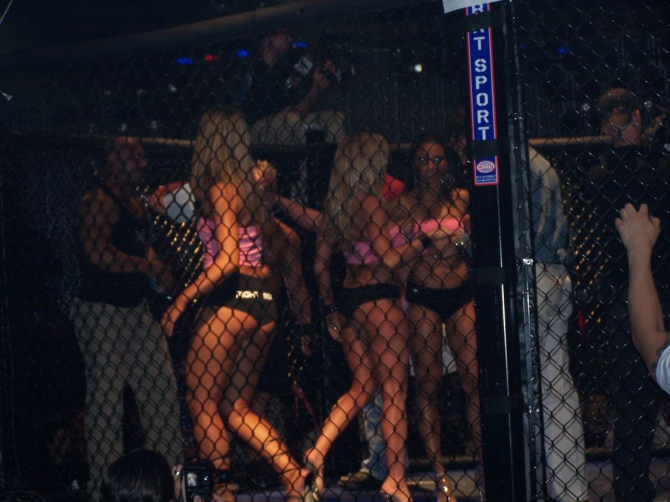 Ring girls in bikinis inside a cage, with spectators behind a fence