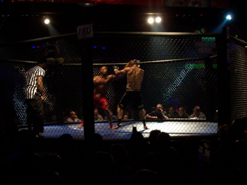 Two fighters in a cage with a referee watching. Spectators are visible in the background