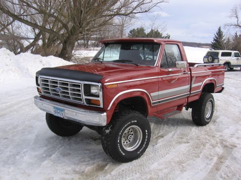 1984 Ford F-150 - This lifted red 1980s Ford F-150 pickup truck is shown in a snowy outdoor setting, with another SUV visible in the background.