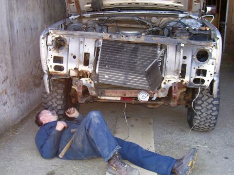 1984 Ford Bronco - A mechanic works on the front of a heavily modified, rusty vehicle, with its hood and grille removed.