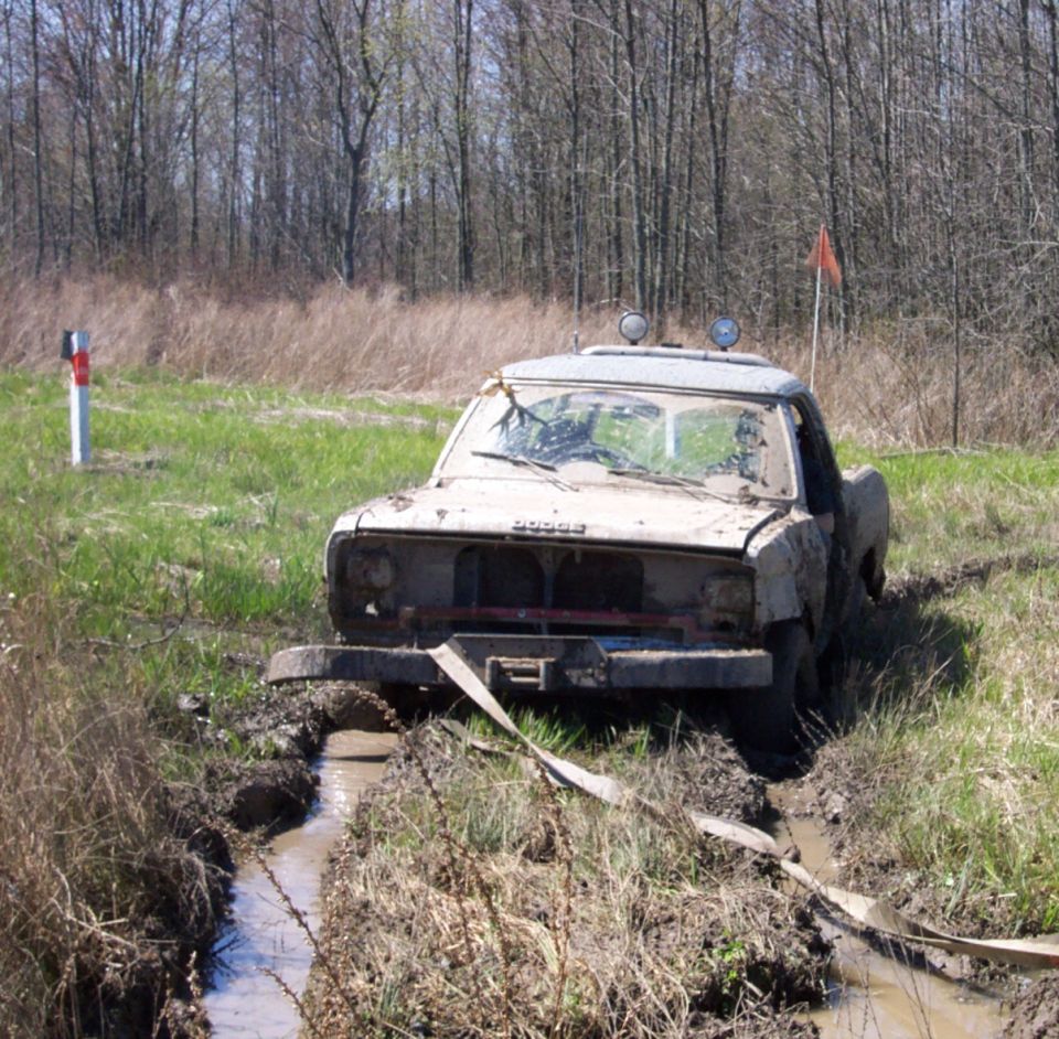Mud-covered Dodge pickup truck with a tow strap, stuck in a muddy ditch with trees in the background