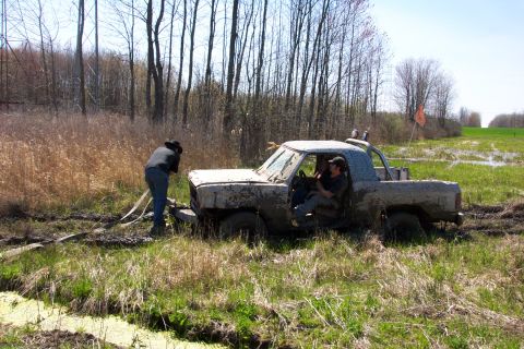 1979 Ford F-150 - Pickup truck stuck deep in a swampy area with two people inside and one person trying to pull it out.
