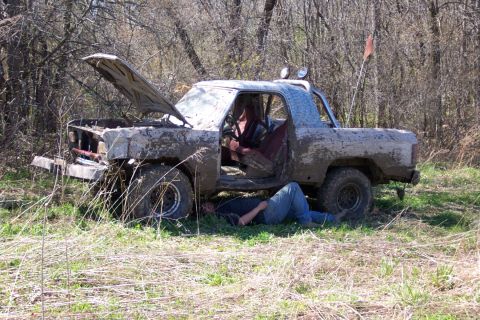 1979 Ford F-150 - A person works on a heavily muddied pickup truck with its hood up, parked in a grassy, wooded area.