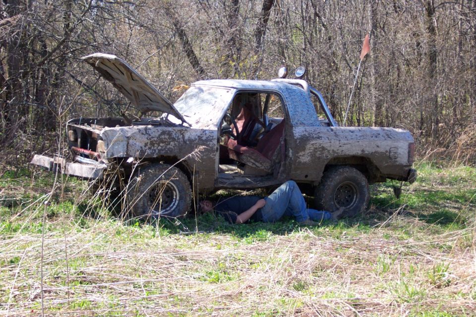 Mud-covered pickup truck with hood open, person working underneath, in a wooded area
