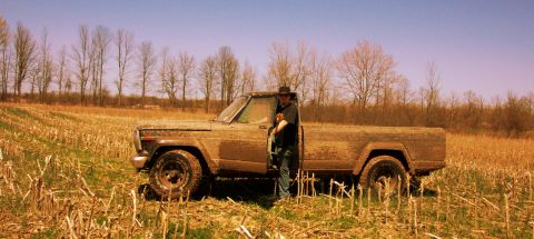 1979 Ford F-150 - A person stands next to a heavily mud-splattered pickup truck in a field of dry stalks under a clear blue sky.