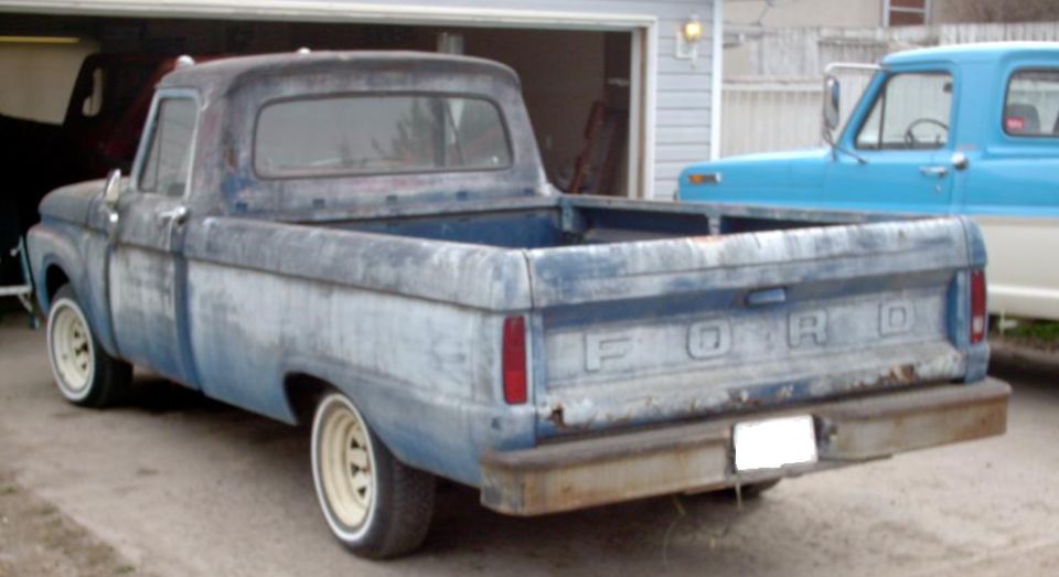 Rear view of a weathered blue Ford pickup truck parked in a driveway, with a blue Ford truck behind it