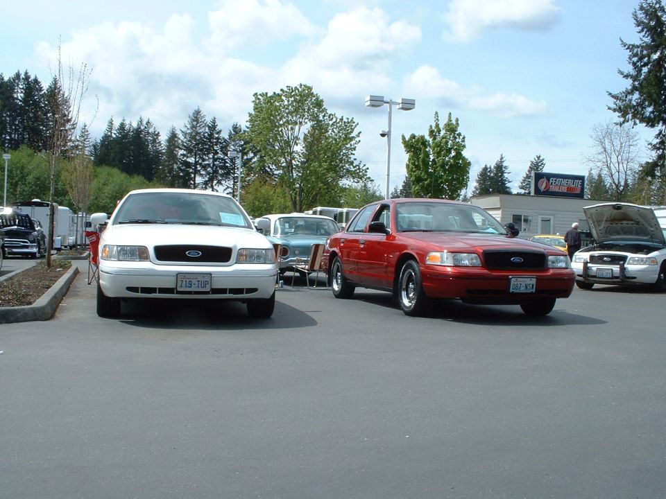 Red Ford Crown Victoria parked on asphalt, front 3/4 view. Hood of a white Crown Victoria open in background