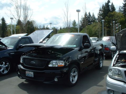 1999 Ford Crown Victoria - A black Ford F-150 SVT Lightning pickup truck is displayed with its hood open at an outdoor event, alongside other vehicles.