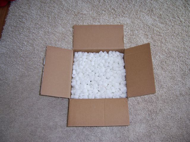 open cardboard box filled with white packing peanuts on a carpeted floor. Supermotors.net logo in corner