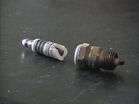 1994 Ford Bronco - A close-up view of two spark plugs, one of which shows damage to its ceramic insulator, resting on a dark, textured surface.