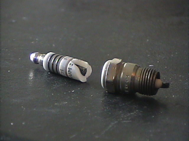 Two spark plugs, one with a broken ceramic insulator, on a dark surface
