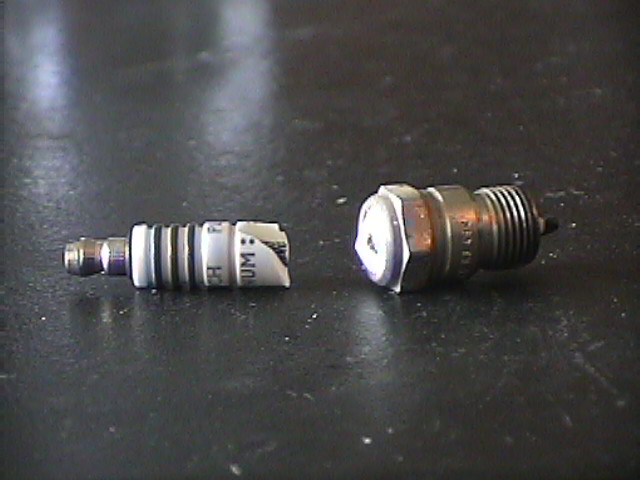 Two spark plugs on a dark surface. One is white with black bands and text, the other is metallic with a white top