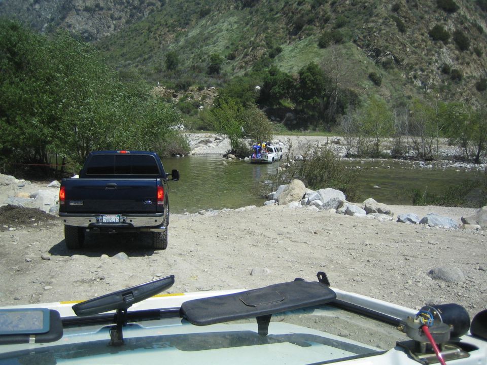 Dark blue Ford F-Series truck parked on dirt near a river, with a white SUV crossing the water in the background