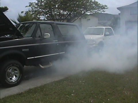 1994 Ford Bronco - A black 1994 Ford Bronco is seen with its hood open and emitting smoke, with a white SUV behind it.