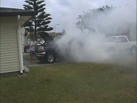 1994 Ford Bronco - A black 1994 Ford Bronco is seen with its hood open and billowing smoke, while a white pickup truck is visible in the background.