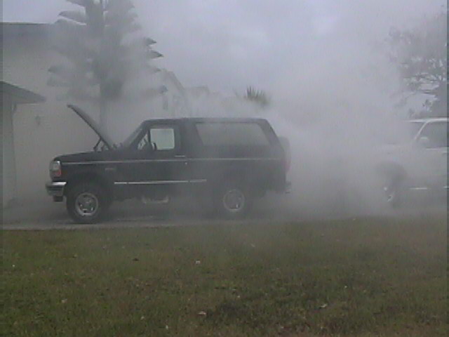 Black 1994 Ford Bronco with hood open, emitting a lot of smoke. Another vehicle is partially visible to the right, also in smoke