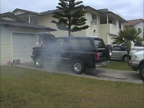 1994 Ford Bronco - A black SUV is emitting a large amount of smoke from its open hood, with a person visible inside.