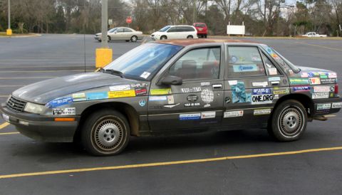 1988 Ford E250 - This heavily stickered sedan, likely from the late 1980s, is parked in a lot with other vehicles visible in the background.