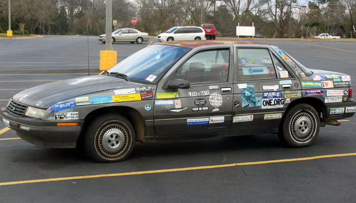 Gray Chevrolet Corsica sedan covered in bumper stickers, driver-side view in parking lot