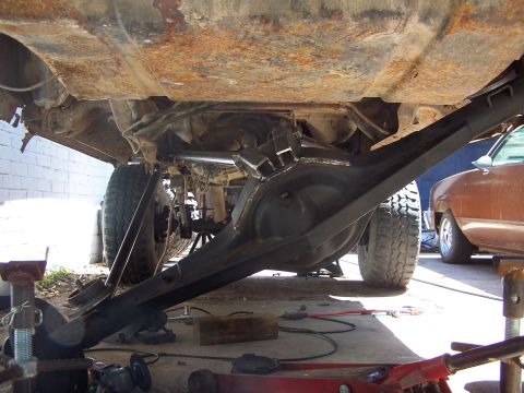 1987 Ford Bronco - Close-up view of the heavily modified undercarriage and rear axle of a lifted vehicle, with another car visible in the background.