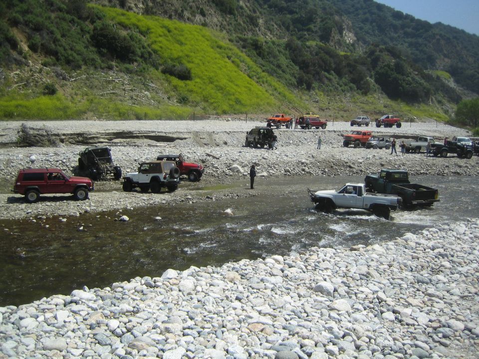 Multiple off-road vehicles, including trucks and SUVs, navigating a shallow, rocky river. Hills with green vegetation rise in the background