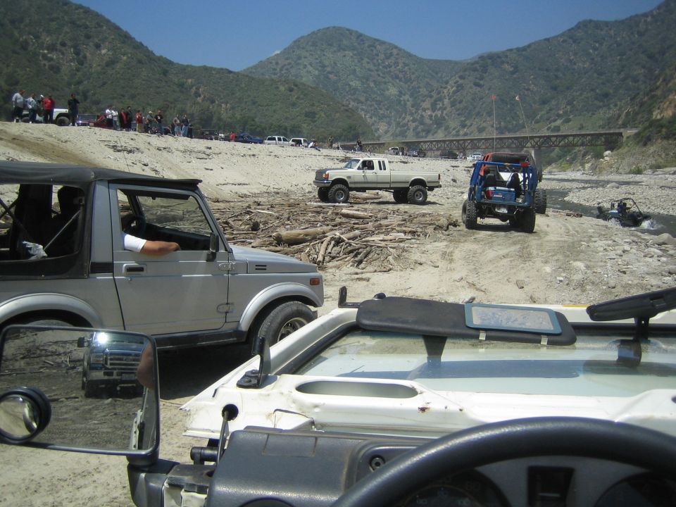 Silver 1987 Suzuki Samurai parked in a dry riverbed with other off-road vehicles and people