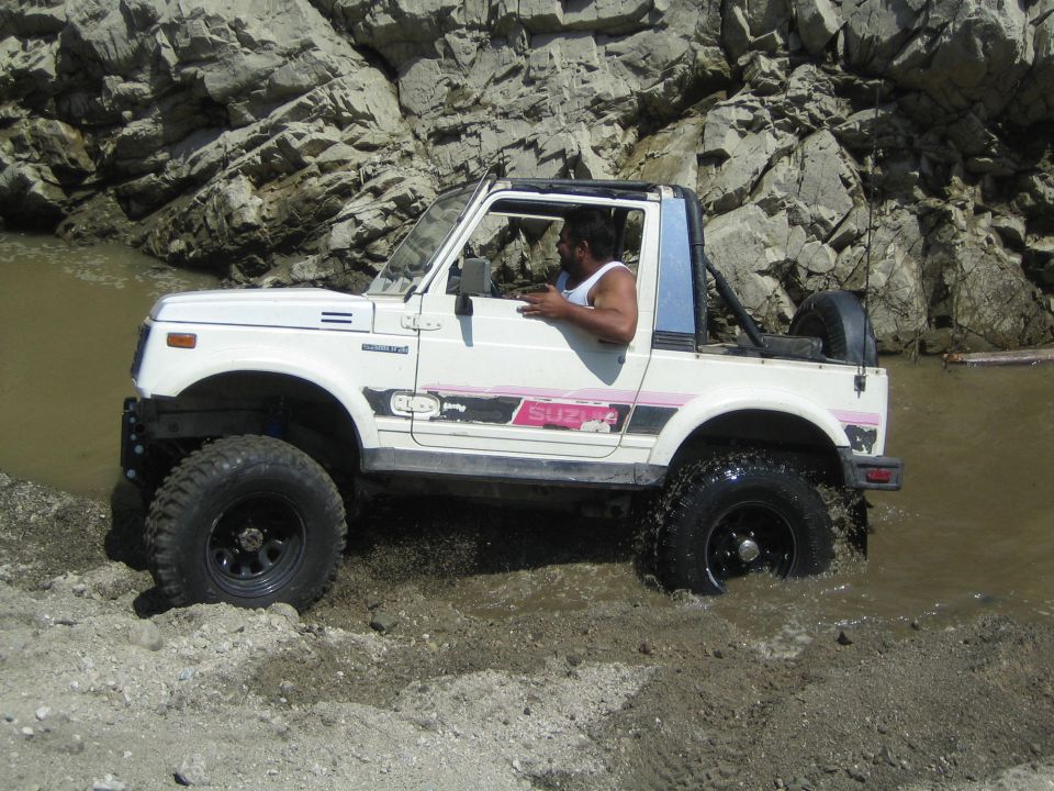 White 1987 Suzuki Samurai with pink and black decals driving through muddy water, side view