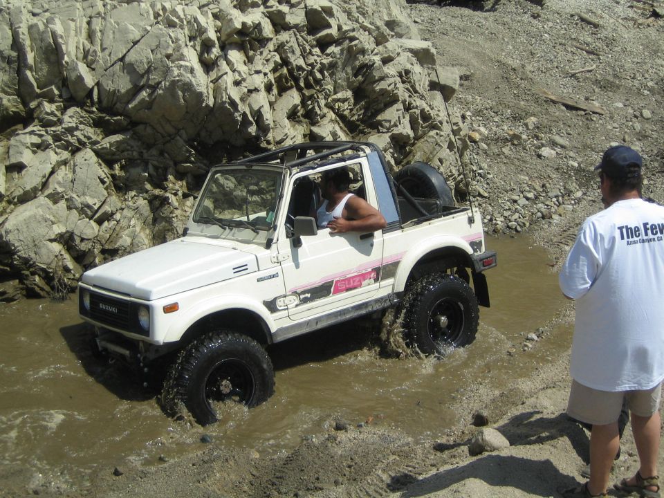 White 1987 Suzuki Samurai driving through water, with a person watching