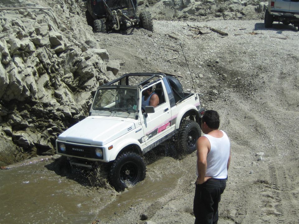 White 1987 Suzuki Samurai driving through water on a rocky trail. Another off-road vehicle is visible behind it