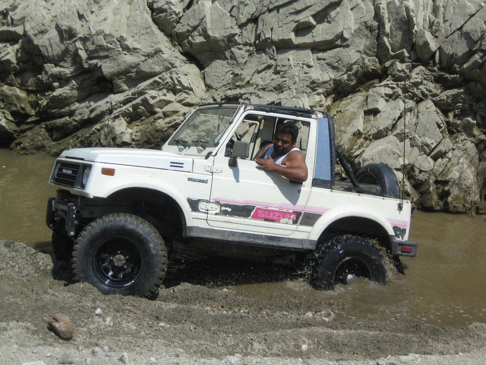 White 1987 Suzuki Samurai with pink stripe driving through shallow water, rocky background