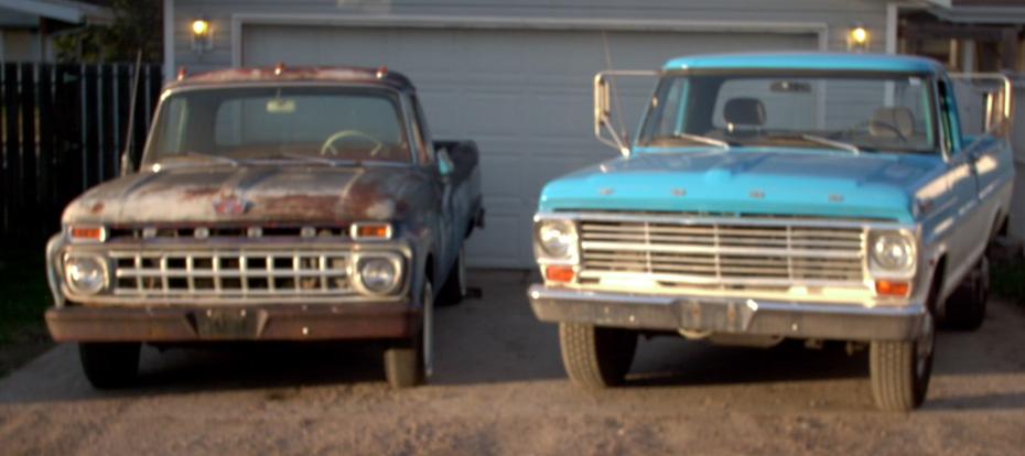 Two old Ford pickups front view, one rusty patina, one turquoise, parked by a garage.