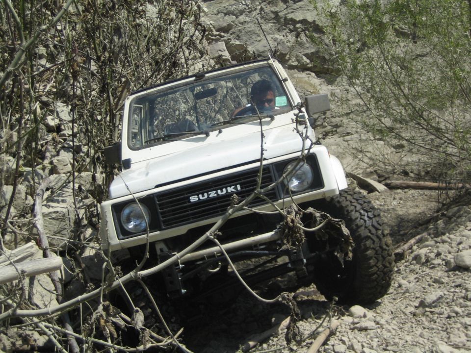 White 1987 Suzuki Samurai navigating a rocky trail with dense brush on the left