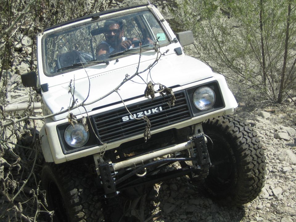 White 1987 Suzuki Samurai driving on a rocky, off-road trail with brush and trees