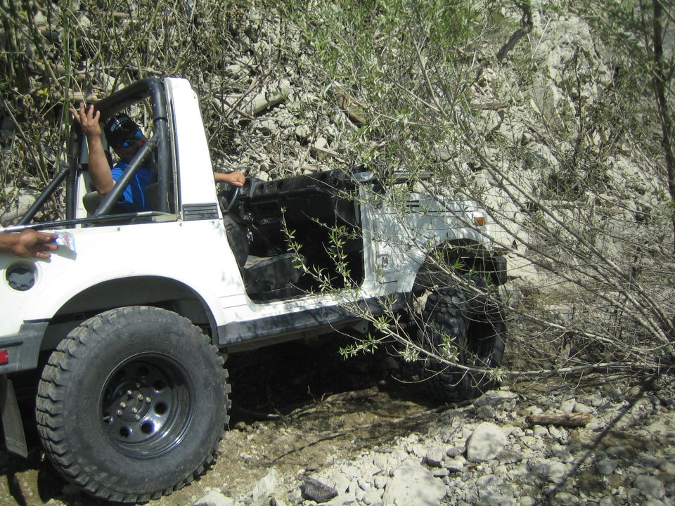 White 1987 Suzuki Samurai off-roading on a rocky, dry creek bed, with the driver's side door removed