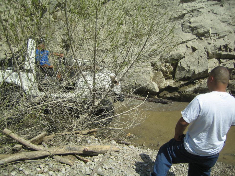 Off-road vehicle partially submerged in muddy water, surrounded by trees and rocks. Person in white shirt watches