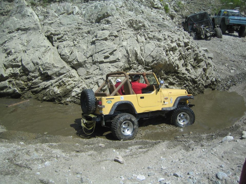 Yellow Jeep Wrangler driving through mud, spare tire on rear, roll cage, rocky cliffside