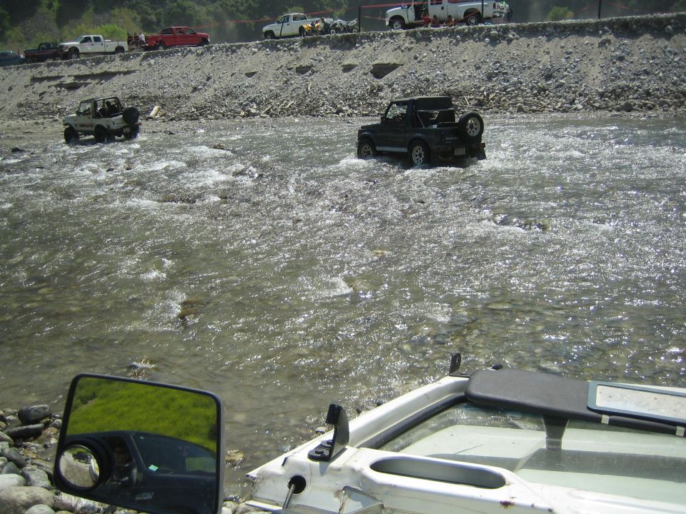 Black Suzuki Samurai driving through a river, with another white Samurai on the bank