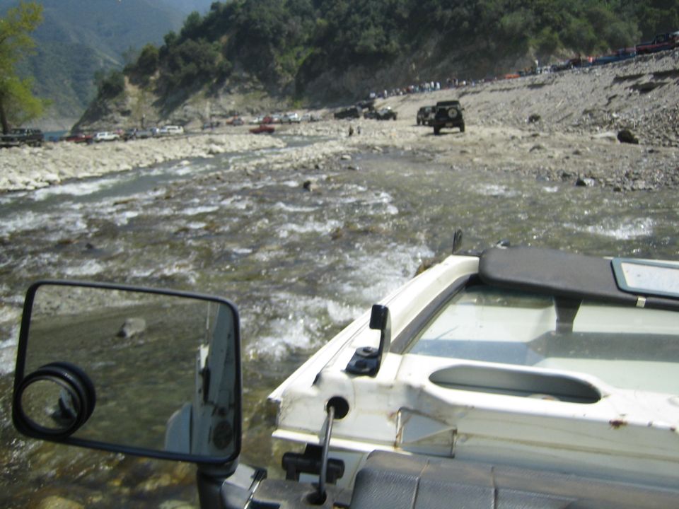 View from inside a white off-road vehicle crossing a shallow, rocky river. Other off-road vehicles are visible on the far bank and in the water