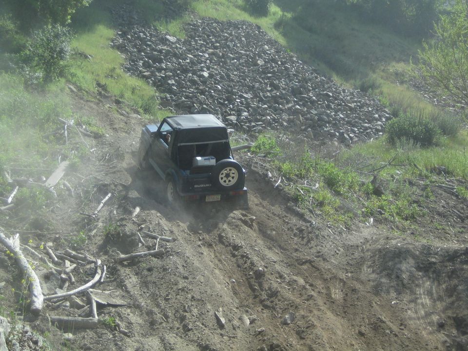 1987 Suzuki Samurai driving up a dusty, steep dirt hill with a pile of rocks in the background
