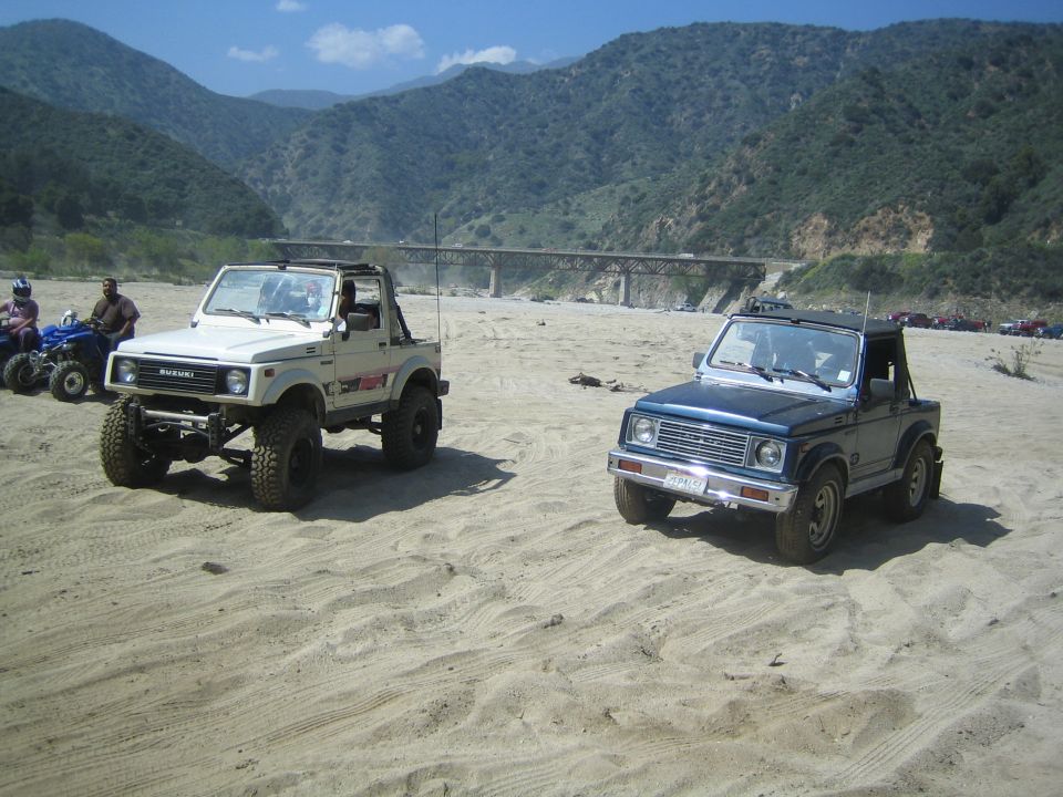 Two Suzuki Samurai SUVs parked on sand with mountains and a bridge in the background. One is white, the other is blue