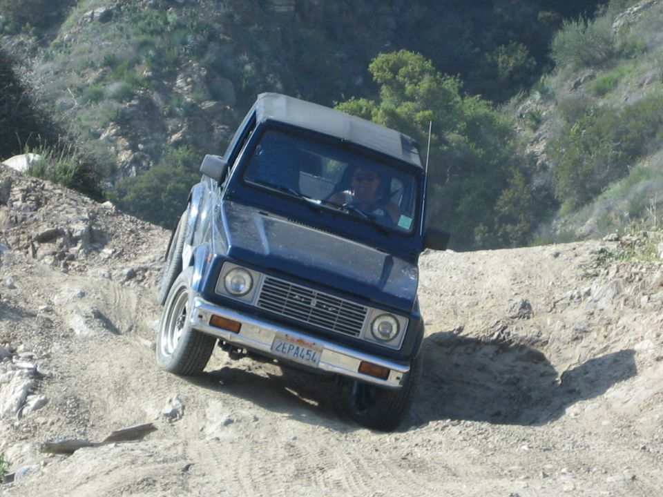 Blue 1987 Suzuki Samurai off-roading on a dirt path, front 3/4 view