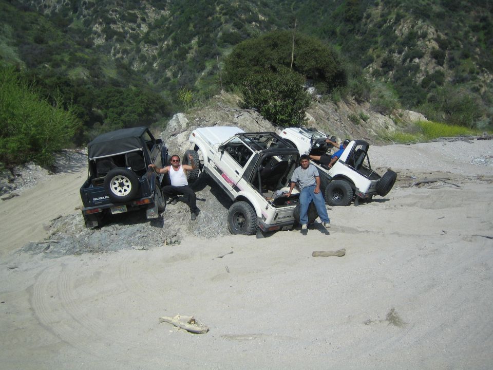 Three Suzuki Samurai off-road vehicles parked on a sandy slope with people sitting on them. One is black, two are white