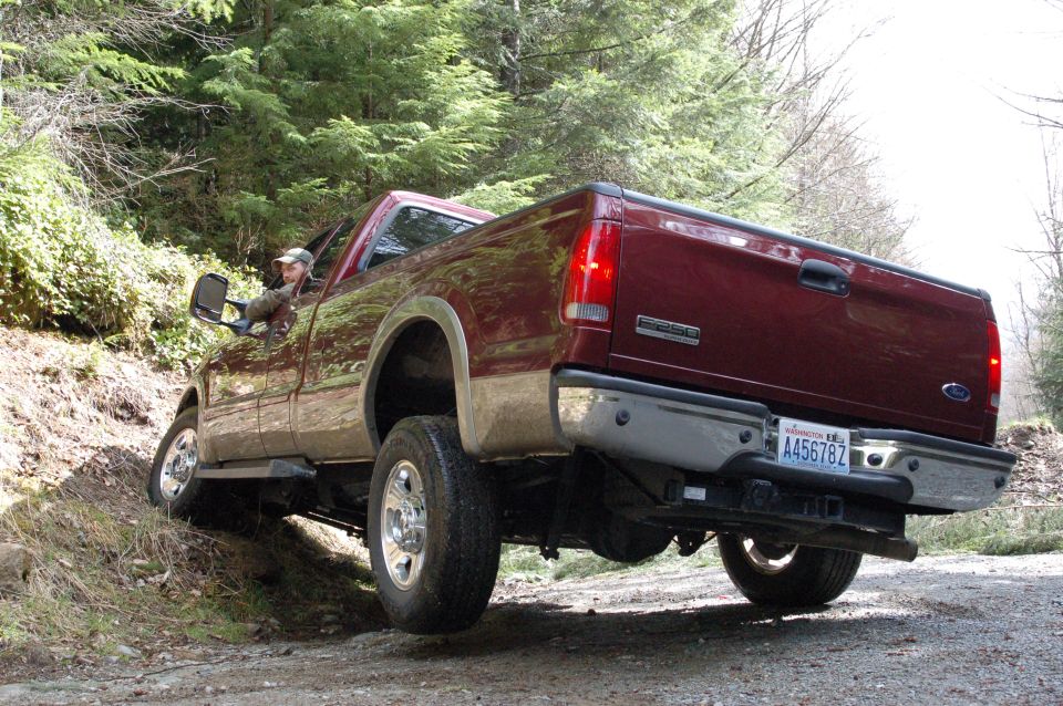 Maroon Ford F-250 Super Duty pickup truck on a dirt road, driver visible, rear 3/4 view