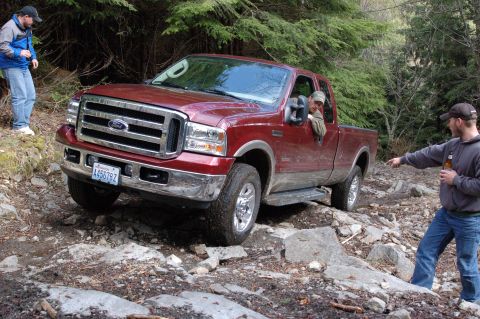 2006 Ford F-250 - A red Ford Super Duty pickup truck is shown driving on a rough, rocky terrain surrounded by trees.