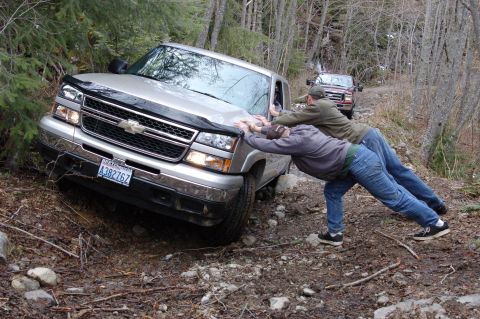 2006 Ford F-250 - Two men push a stuck silver Chevrolet Silverado truck on a muddy, wooded trail while another truck follows behind.