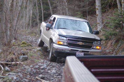 2006 Ford F-250 - A silver pickup truck navigates a challenging, rocky trail through a wooded area.